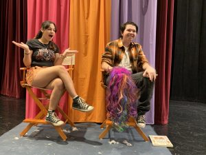 Two students from West Chicago Community High School sit in director's chairs on a stage. A multi-colored backdrop appears behind them.