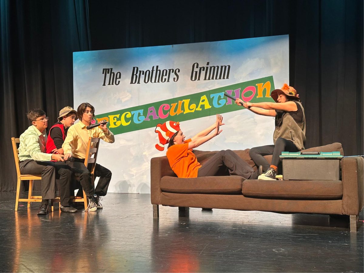The Brothers Grimm Spectaculathon sign hangs in the background while students battle each other on a brown sofa on stage.