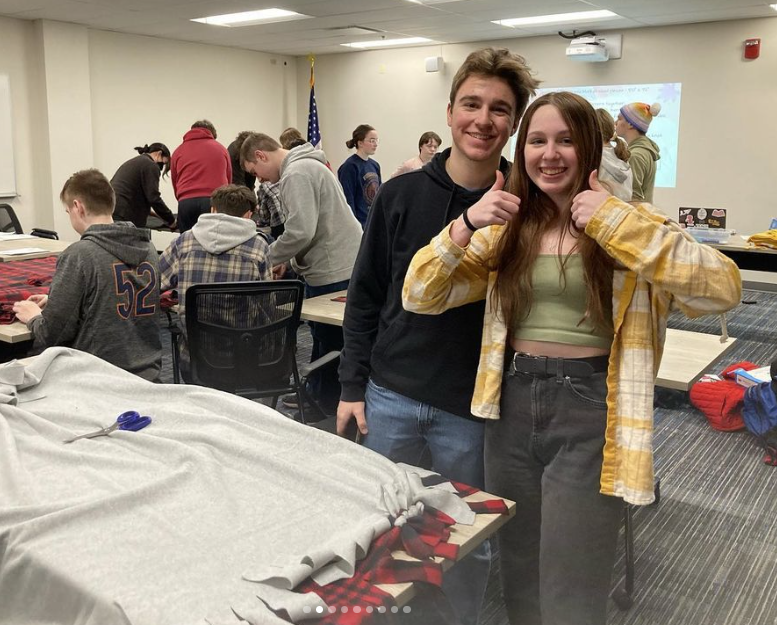 Two students, a male and a female, stand behind a fleece blanket, half-constructed. Other students work on similar tie-blanket projects in the background.