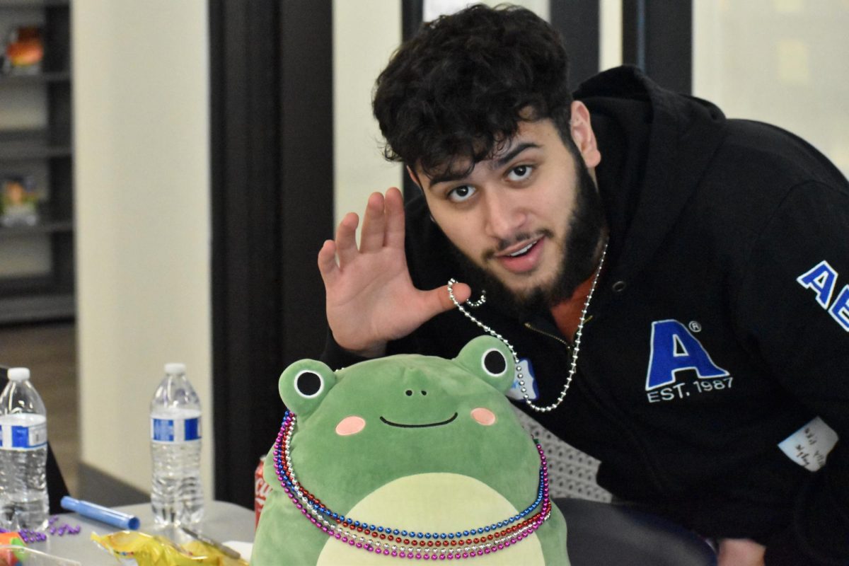 Senior Qssam Alwan poses with a frog  squishmallow, a support stuffed animal. All while showing off his Group 1 accessory, a beaded necklace. 