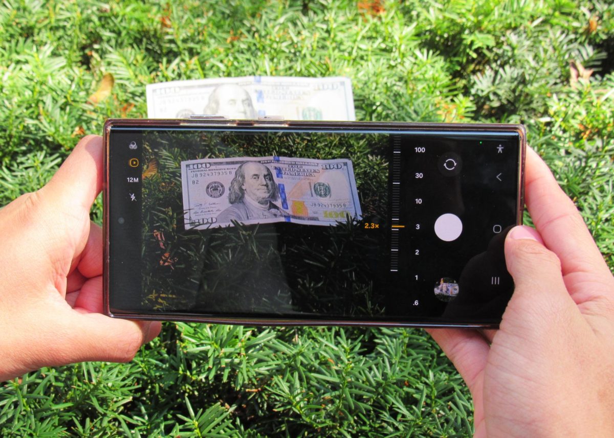 Un estudiante fotografía un billete de $100 parcialmente escondido entre arbustos durante la creación de la exhibición “That’s Why I’m Here”. El maestro de ESL Mark Poulterer dijo que la imagen refleja cómo las oportunidades económicas en los Estados Unidos pueden parecer visibles, pero fuera del alcance para muchas familias inmigrantes que equilibran trabajos de bajos salarios y gastos diarios. (Foto proporcionada por Jaime, apellido reservado)